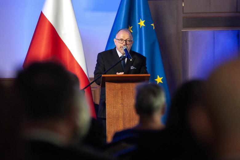 Sławomir Strzelecki wypowiada się z podium w stronę publiczności. W tle flagi Polski i Unii Europejskiej