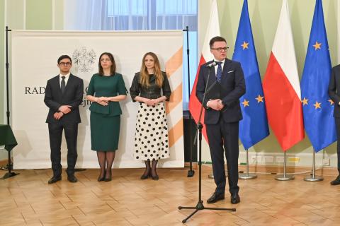 Osoba przemawia do mikrofonu na tle flag Polski i UE, za nią trzy osoby stoją i słuchają wystąpienia