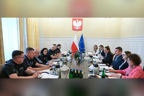 Uczestnicy spotkania - szesnaście osób - rozmawiają przy stole konferencyjnym. W szczycie stołu znajdują sie godło Polski oraz flagi Polski i Unii Europejskiej