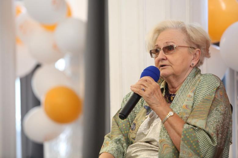 zdjęcie: kobieta w okularach siedzi i mówi do mikrofonu, za nią widać pomarańczowo-białe balony
