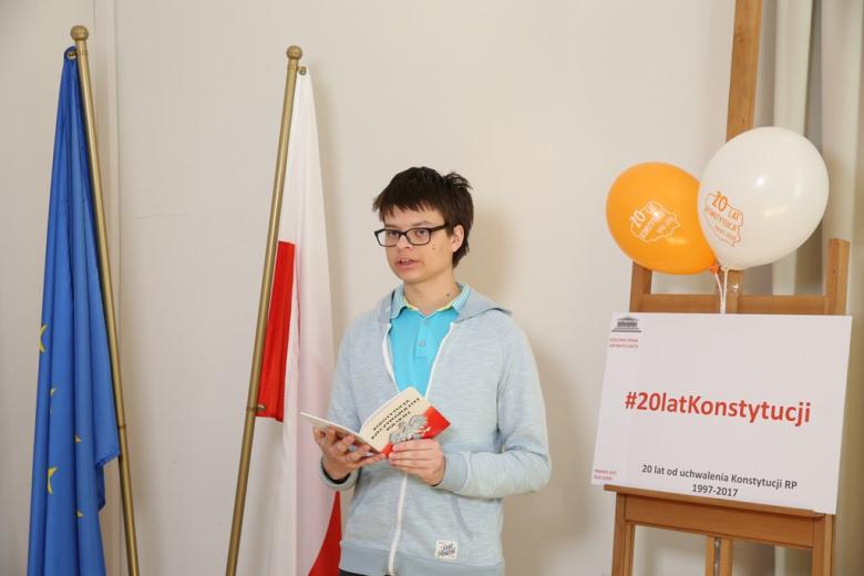 zdjęcie: młody chłopak stoi przy polskiej i unijnej fladze i trzyma w ręku Konstytucję