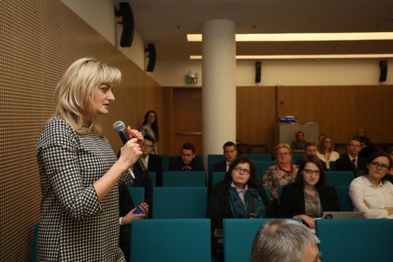 zdjęcie: kilka osob siedzi na sali, jedna z kobiet stoi i mówi do mikrofonu