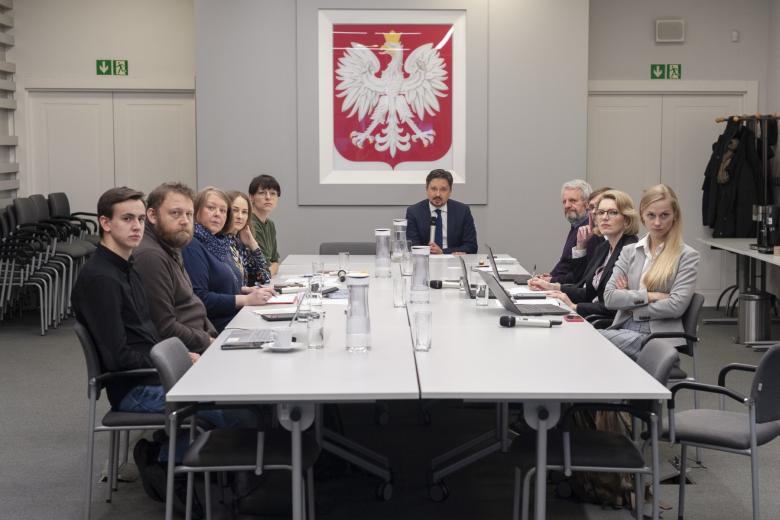 Grupa kilkunastu osób siedząca po obu stronach stołu konferencyjnego, w tle duże godło Polski