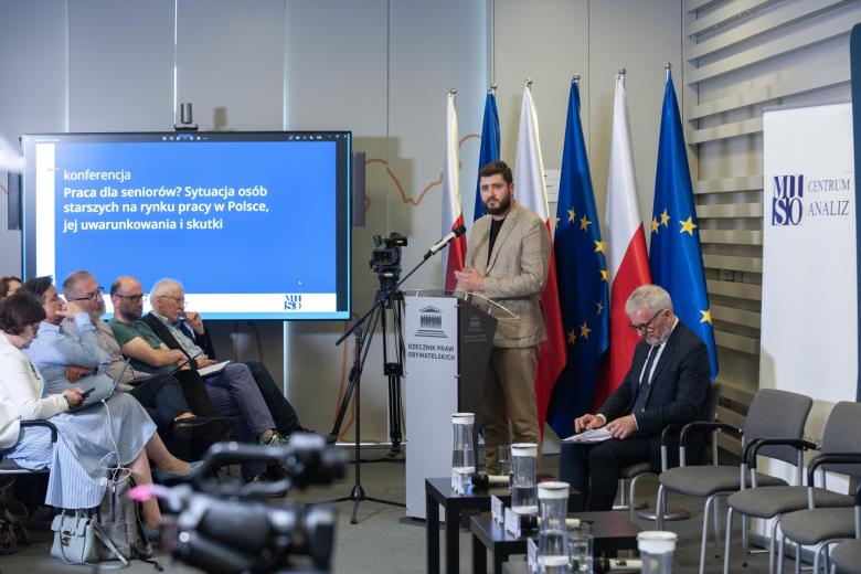 Mateusz Komorowski przemawia za mównicą na tle flag Polski i UE