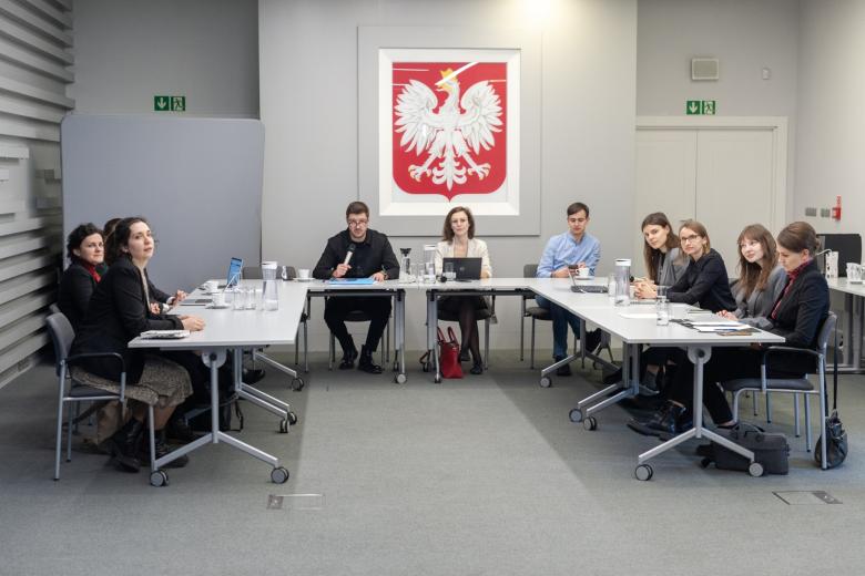 Kilkanaście osób siedzi przy stole konferencyjnym i rozmawia, w tle godło Polski z białym orłem na czerwonym tle