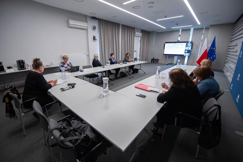 W sali konferencyjnej członkowie komisji siedzą przy ustawionym w kształt podkowy konferencyjnym stole i oglądają prezentację wyświetlaną na ekranie znajdującym się w szczycie stołu konferencyjnego. Na ekranie wyświetleni są również członkowie Komisji biorący udział w spotkaniu za pośrednictwem Internetu. W tle flagi Polski i Unii Europejskiej