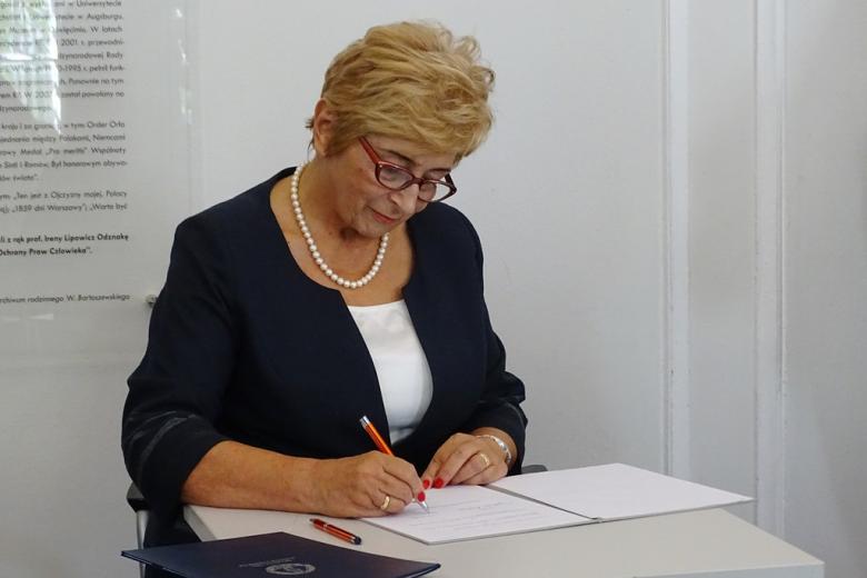 zdjęcie: kobieta w blond włosach siedzi przy białym stoliku i podpisuje dokument