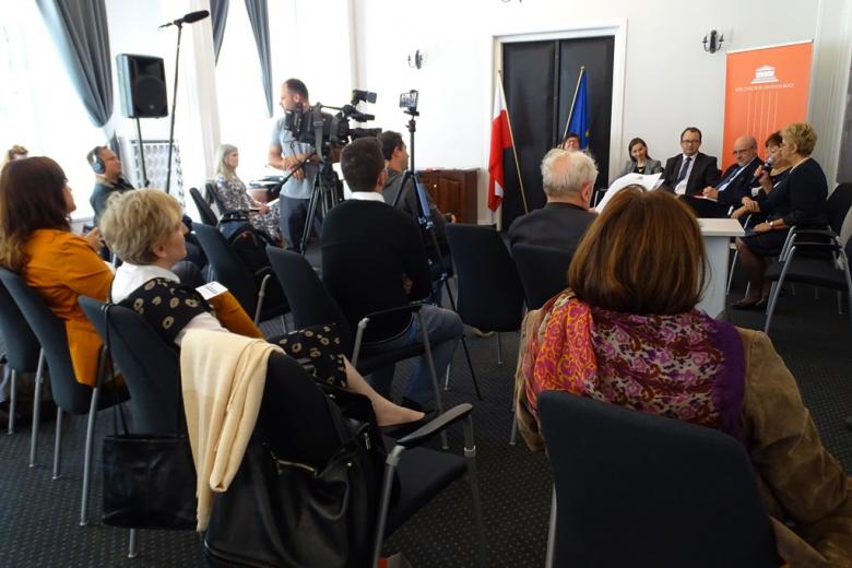 zdjęcie: kilka osób siedzi na sali, widoczna jest kamera i mikrofony