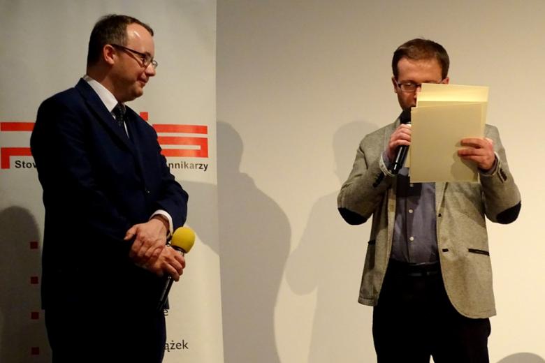 na zdjęciu stoją dwaj mężczyźni, mężczyzna po prawej stronie trzyma w ręku kartki, z ktorych odczytuje fragmenty tekstu