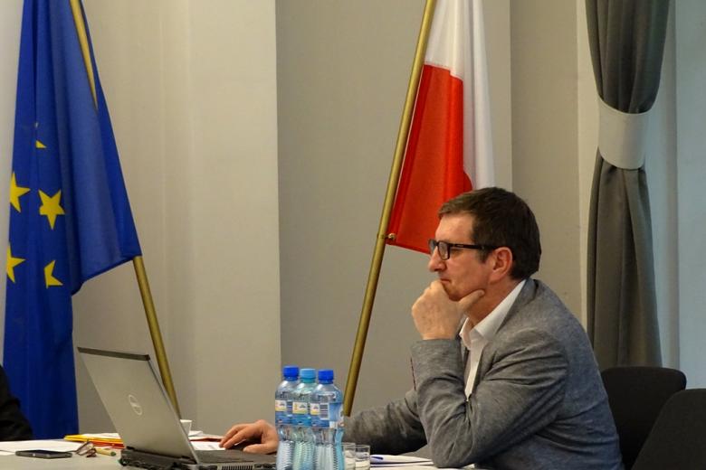 zdjęcie: mężczyzna siedzi prz białych stołach, za nim flagi Polski i UE