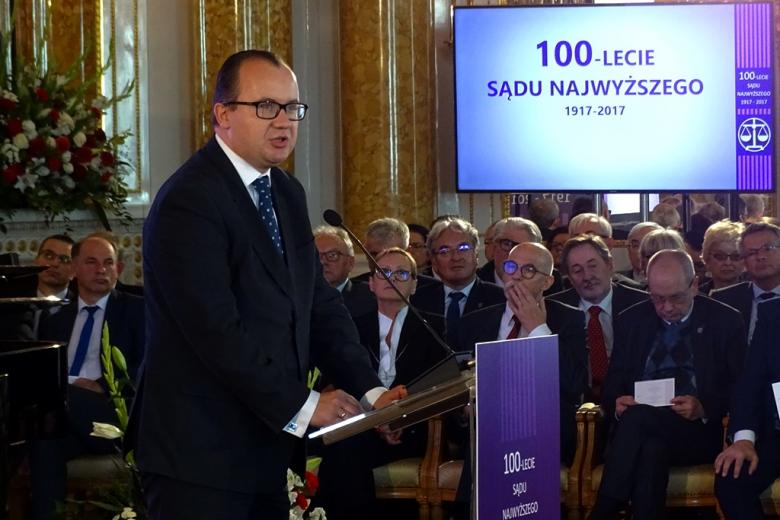 zdjęcie: mężczyzna w garniturze stoi przy mównicy za nim siedzi kilkanaście osób