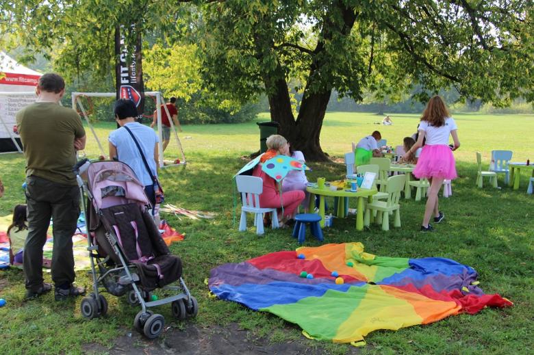 zdjęcie: na trawie lezy kolowory koc, w tle widać małe kolorowe krzestałka i stoliki przy których siedzą dzieci