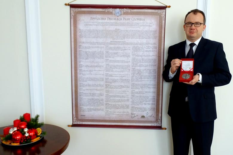 zdjęcie: mężczyzna w garniturze stoi po prawej stronie i trzyma w dłoniach pudełko ze srebrnym medalem, za nim wisi tekst Powszechnej Deklaracji Praw Człowieka, a po lewej stronie widać stolik ze świątecznym stroikiem