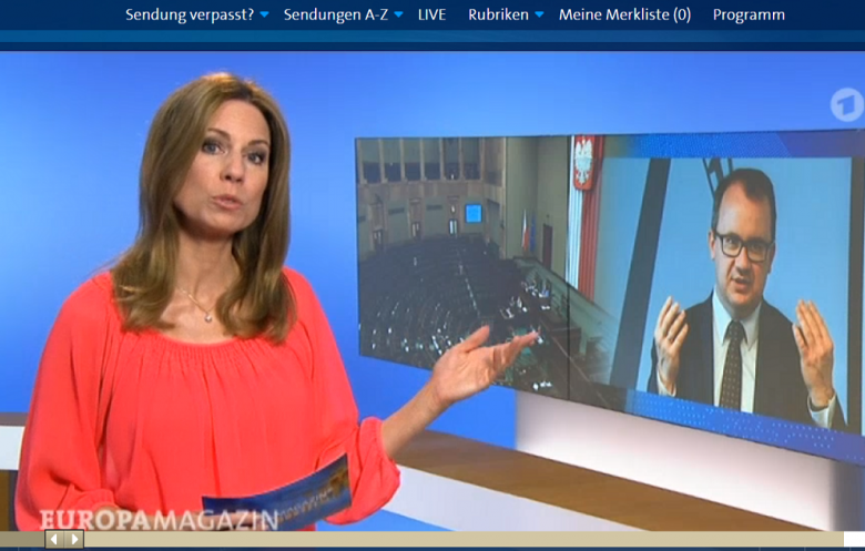 Screen programu telewizyjnego - kobieta w czerwonym pokazuje na ekran, na którym widać mężczyznę w garniturze