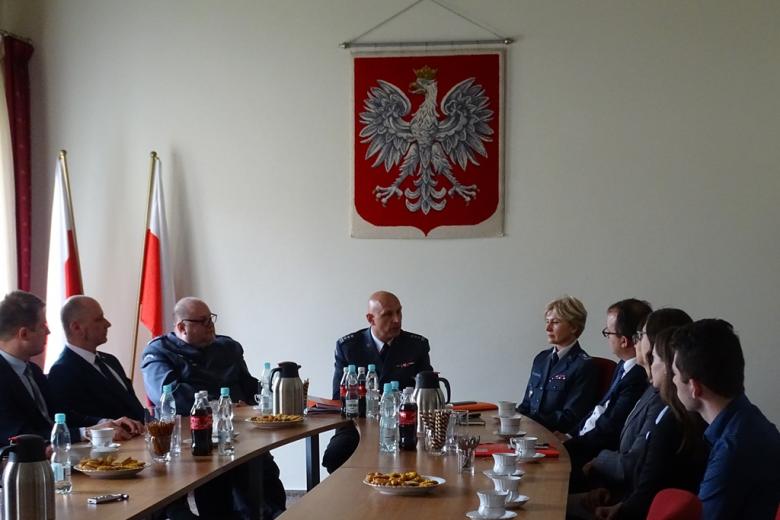 Zdjęcie: ludzie przy stole, trójka w mundurach, z tyłu dwie biało-czerwone flagi i duże godło