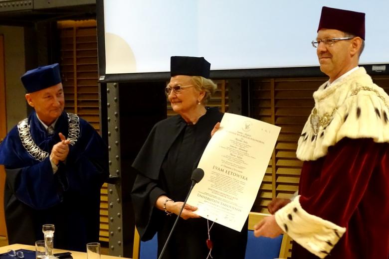 zdjęcie: kobieta w czarnej todze pokazuje dyplom po jej obu stronach stoją mężczyźni w togach