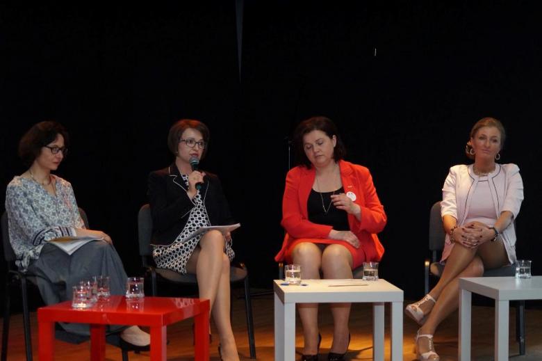 Cztery kobiety (w szarym, czarnym, czerwonym i białym stroku) dyskutują w panelu