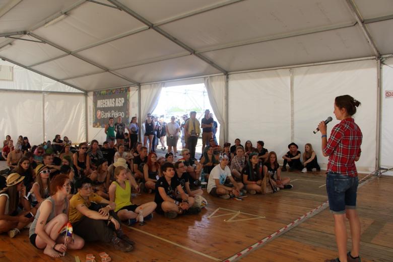 zdjęcie: duża grupa ludzi siedzi na podłodze w dużym namiocie, na pierwszym planie kobieta stoi i mówi do mikrofonu