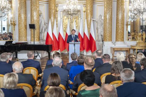 RPO Marcin Wiącek przemawia za mównicą na tle ośmiu polskich flag w dużej sali w Zamku Królewskim w Warszawie