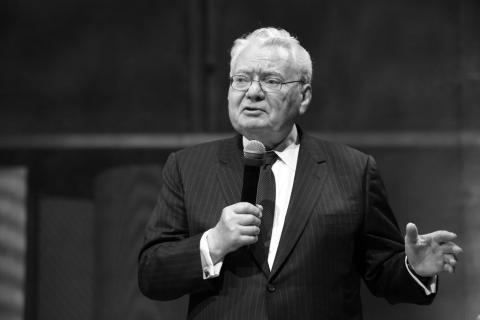 Czarno białe zdjęcie przedstawiające mężczyznę w garniturze przemawiającego do mikrofonu trzymanego w ręce