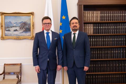 Dwaj mężczyźni w garniturach pozują do wspólnego zdjęcia na stojąco na tle flag Polski i UE