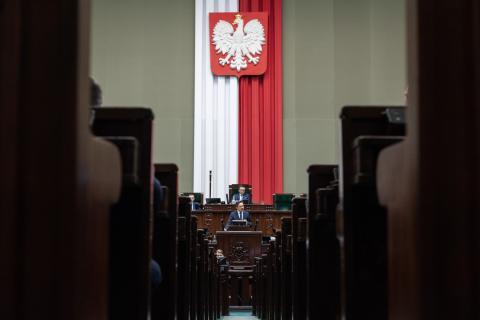 Osoba przemawia za mównicą na sali plenarnej Sejmu RP, w tle na ścianie godło Polski - biały orzeł w koronie na czerwonym tle