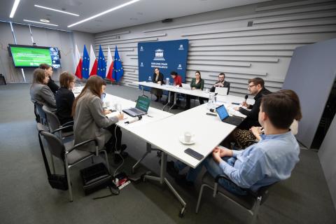 Kilkanaście osób siedzi przy stole konferencyjnym i rozmawia, w tle flagi Polski i UE