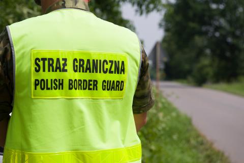 Strażnik graniczny stojący tyłem do kadru przy leśnej drodze. Strażnik ma na sobie kamizelkę odblaskową z napisem "Straż Graniczna - Polish Border Guard"