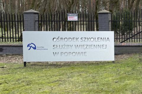 Tablica z napisem "Ośrodek Szkolenia Służby Więziennej w Popowie", w tle ogrodzenie i las