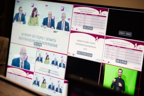 Zdjęcie monitora kontrolnego transmisji konferencji prasowej Państwowej Komisji Wyborczej. Na monitorze widać ujęcia na uczestników konferencji, plansze z elementami infograficznymi i nagrywanego tłumacza polskiego języka migowego.