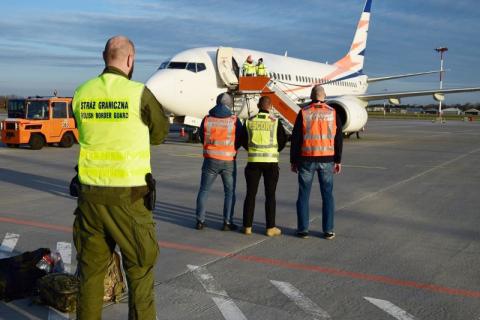 Strażnik graniczny, eskortowana osoba oraz dwóch konwojentów stoją przed samolotem na płycie lotniska
