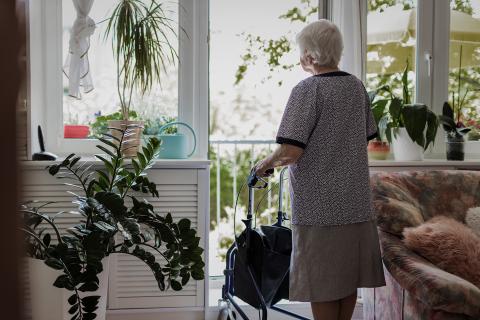 Starsza kobieta z chodzikiem stoi tyłem do kadru i wygląda na balkon w swoim mieszkaniu